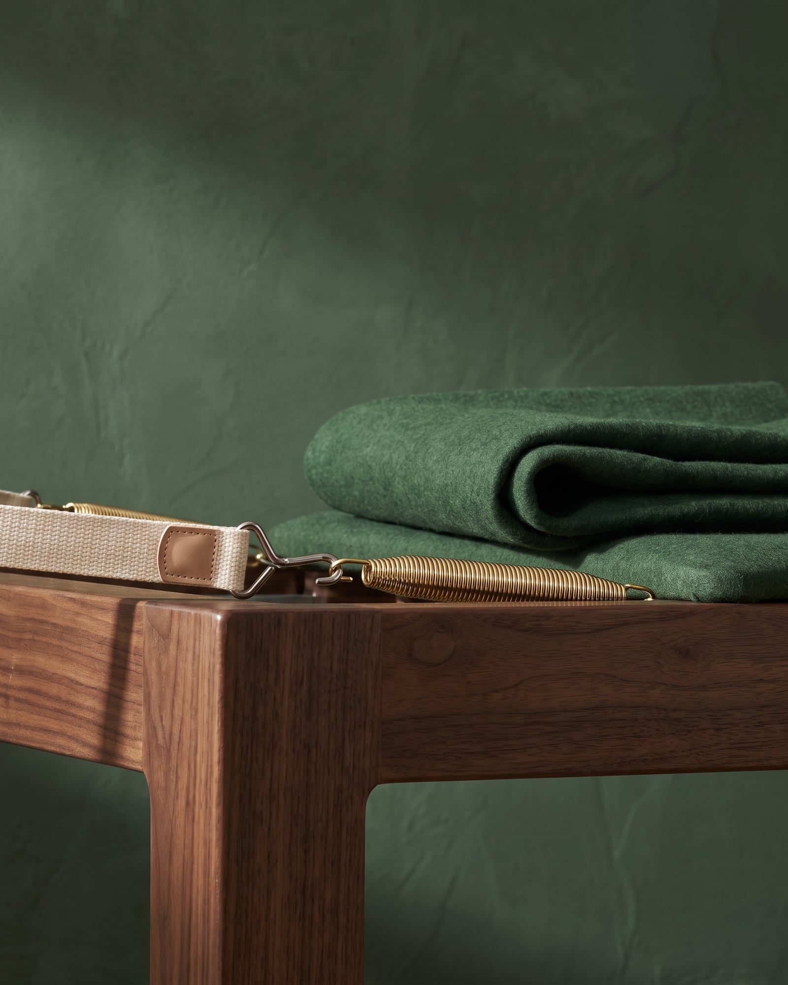 Reformer carriage edge with brass spring, woven ivory strap, and folded green felt against a deep green wall